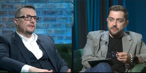 Split-screen interview with two men seated on green couches, one in a dark suit on the left and the other in a gray blazer on the right, talking.