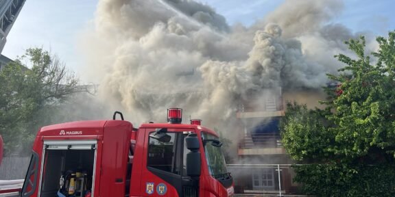 Red fire engine in the foreground as a building emits a thick plume of smoke from a fire behind it.