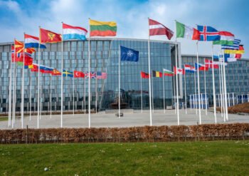 Modern glass-front building with row of flagpoles displaying numerous international flags in front of the plaza under a blue sky